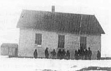 Black and white photo of a schoolhouse with children standing beside it