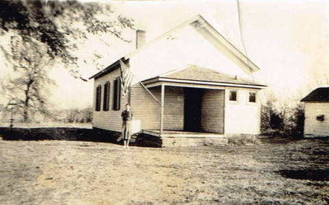 Photo of a white schoolhouse with a teacher