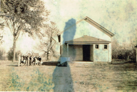 Photo of a white schoolhouse with children saluting a flag outside	