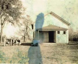 Photo of a white schoolhouse with children saluting a flag outside	