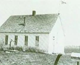 White schoolhouse with a flag, water pump, and chimney